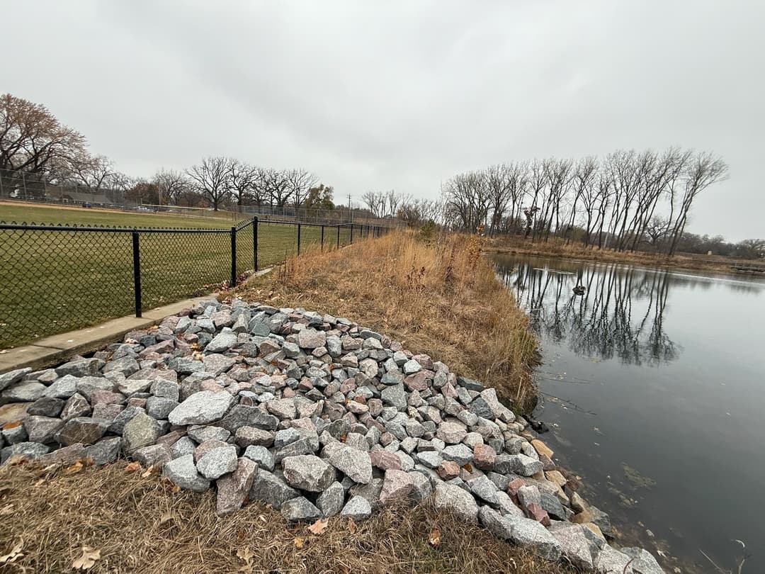 Rocky shoreline along a pond with grass and trees under a cloudy sky.